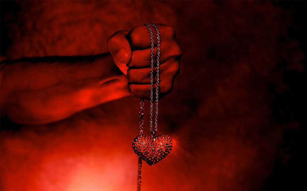 A strong clenched fist holding a sparkling heart-shaped pendant on a chain, illuminated in a deep red glow