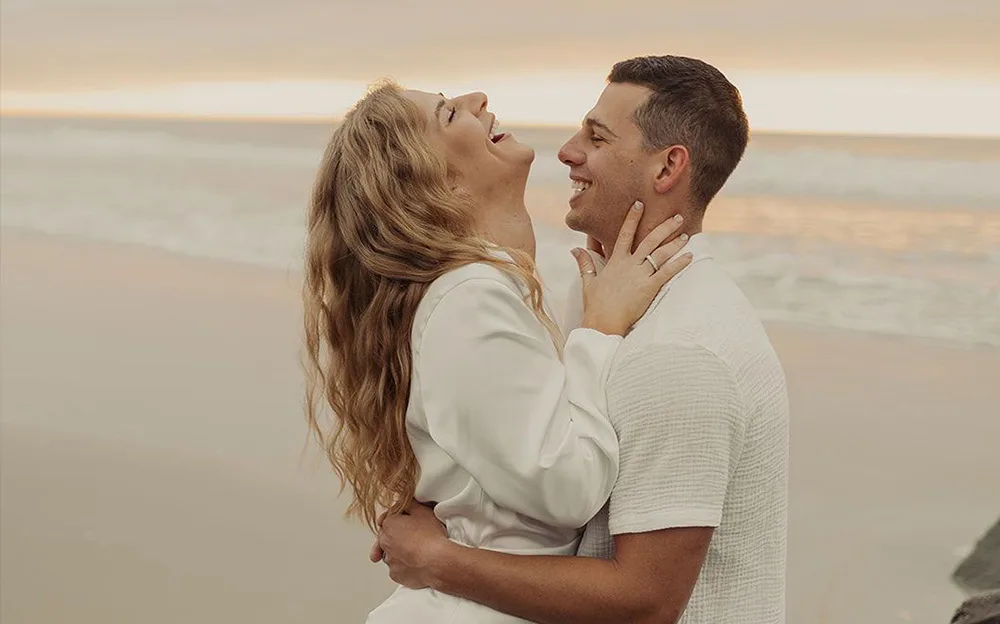 A happy couple embracing and laughing on a beach at sunset, with soft golden hues reflecting on the water