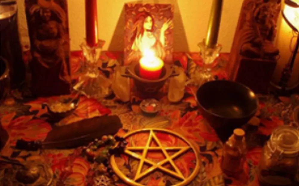 A spiritual altar featuring lit candles, a pentagram, feathers, crystals, and various sacred items arranged on a colorful patterned cloth