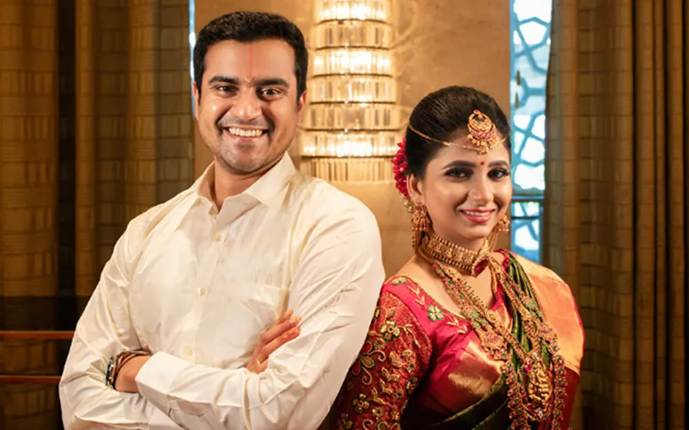 Smiling couple in traditional South Indian wedding attire, adorned with jewelry and vibrant outfits indoors