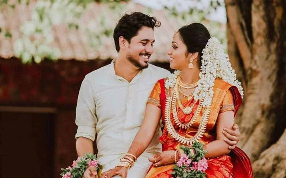 A traditional Indian wedding couple sitting under a tree, dressed in vibrant red and white attire, sharing a joyful moment